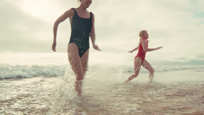 A happy mother and her young daughter joyfully run and splash through the shallow ocean waves together in slow motion