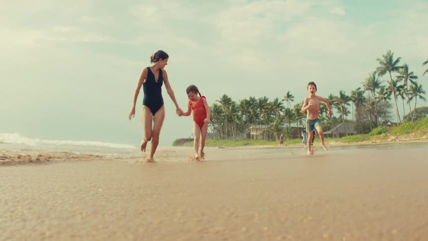 A joyful mother and her two young children hold hands and run along the shoreline of a tropical beach in slow motion