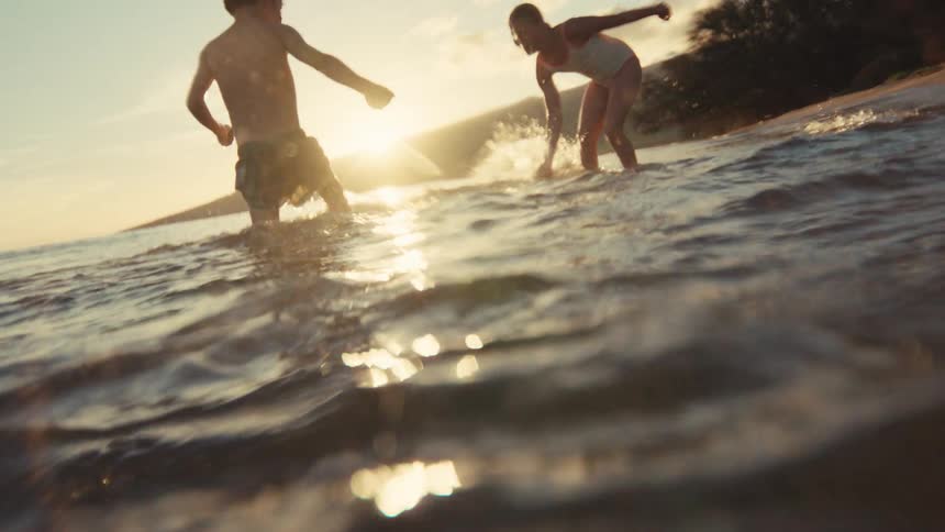 A young boy and girl splash each other and play in the shallow ocean waves at sunset, captured in slow motion from a low, underwater perspective