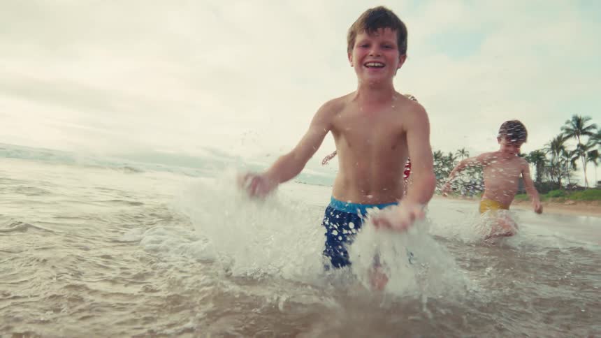 Joyful children laugh as they run and splash in the shallow ocean waves on a tropical beach in slow motion