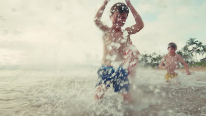 Joyful children laugh as they run and splash in the shallow ocean waves on a tropical beach in slow motion