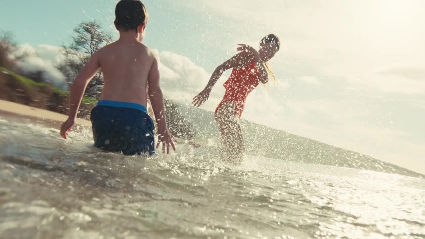 A young boy joyfully splashes his mom in the shallow ocean waves on a sunny day in cinematic slow motion