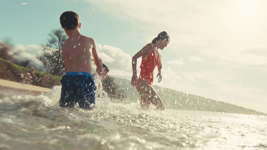 A young boy joyfully splashes his mom in the shallow ocean waves on a sunny day in cinematic slow motion