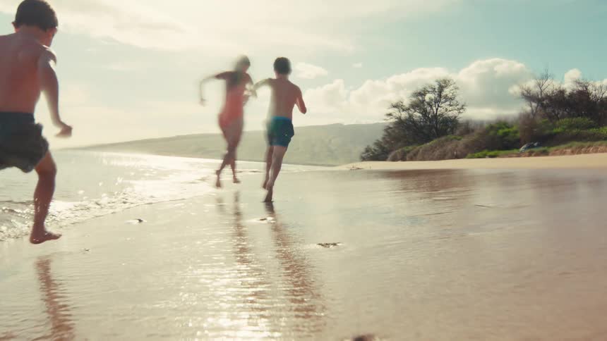 Slow motion shot of two boys and a woman running barefoot along the waters edge on a beautiful tropical beach