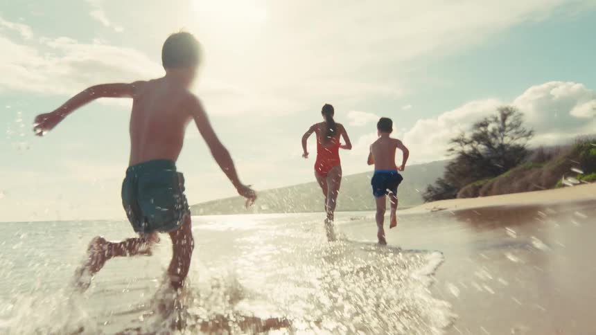 Slow motion shot of two boys and a woman running barefoot along the waters edge on a beautiful tropical beach
