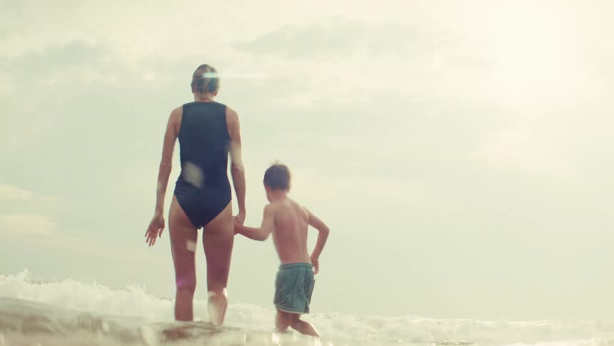 In slow motion, a woman and her young boy hold hands and playfully walk into the sunlit ocean surf