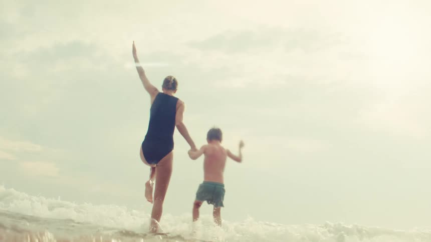 In slow motion, a woman and her young boy hold hands and playfully walk into the sunlit ocean surf