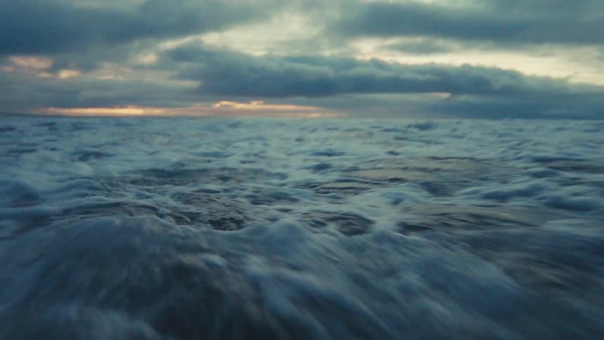 Immersive low-angle slow motion shot of an ocean wave crashing over the camera on a moody, cloudy evening at the beach