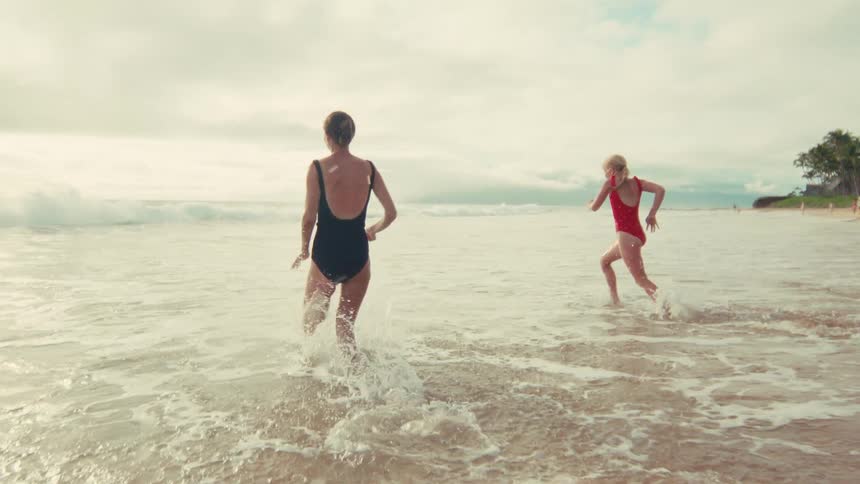 A mother and her young daughter playfully run into the ocean waves together in slow motion on a beautiful beach