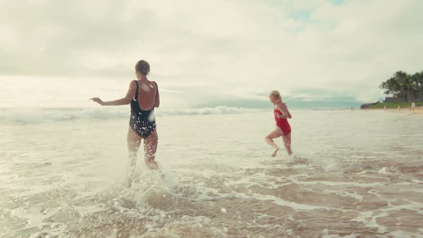 A mother and her young daughter playfully run into the ocean waves together in slow motion on a beautiful beach
