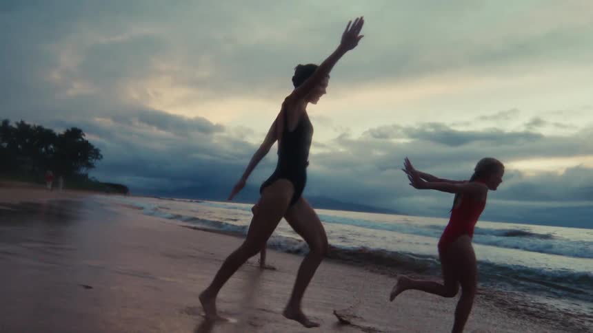 A mother and her two children playfully run and skip along the ocean's edge at sunset in slow motion