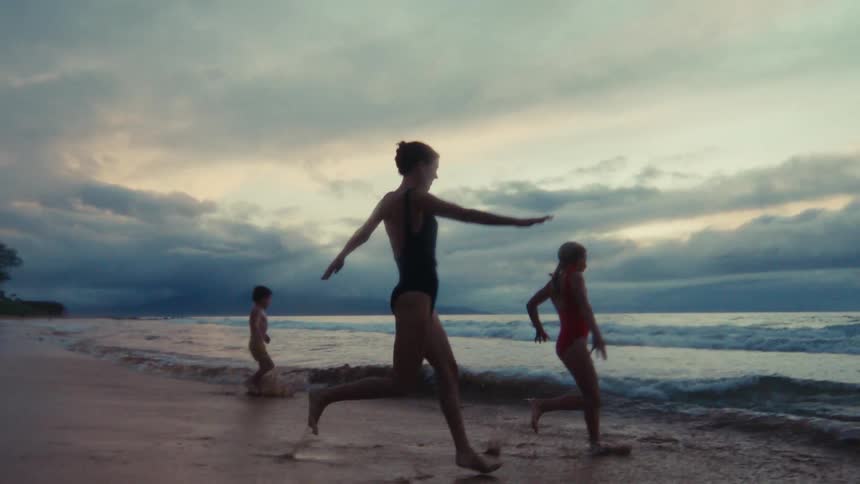 A mother and her two children playfully run and skip along the ocean's edge at sunset in slow motion