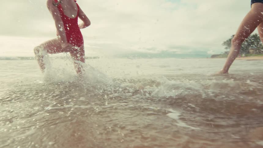 Two joyful young women in swimsuits run and splash through the shallow ocean water on a tropical beach in slow motion
