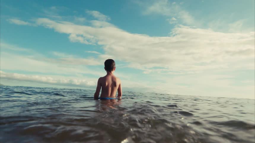 From the water's surface, a young boy contemplatively watches the horizon from the calm ocean in slow motion