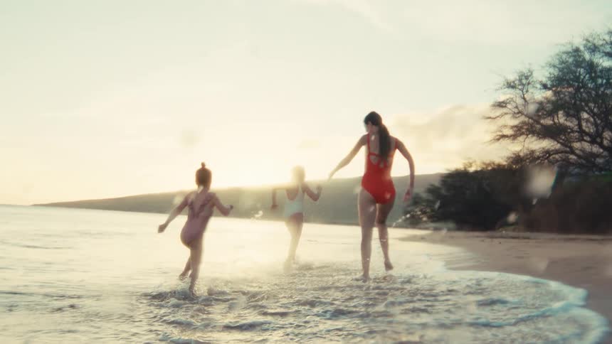 A mother and her two young daughters playfully run and splash in the shallow ocean waves at sunset in slow motion