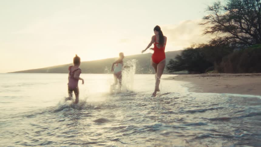 A mother and her two young daughters playfully run and splash in the shallow ocean waves at sunset in slow motion