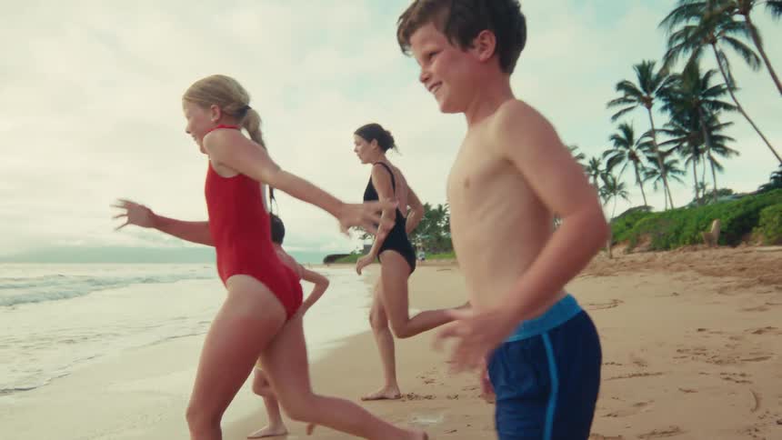 Joyful children and their mother run along the sandy shoreline of a tropical beach in slow motion