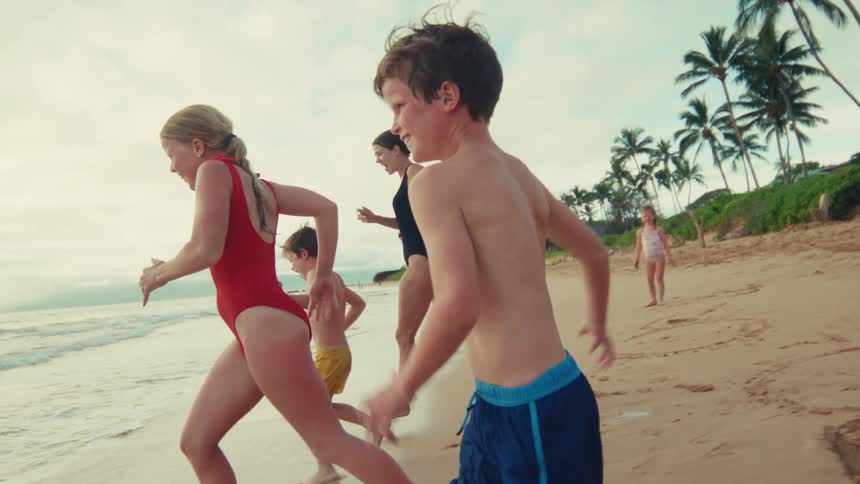 Joyful children and their mother run along the sandy shoreline of a tropical beach in slow motion