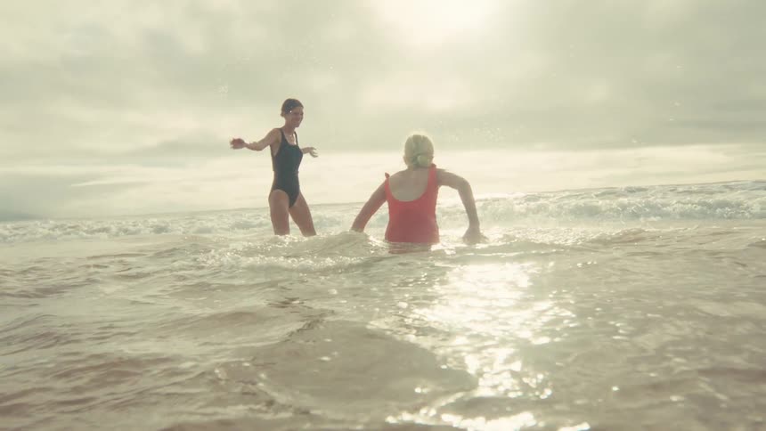 Two playful women in swimsuits enjoy a sunny day at the beach, splashing each other in the shimmering ocean waves in slow motion