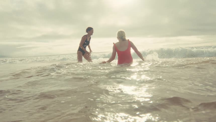 Two playful women in swimsuits enjoy a sunny day at the beach, splashing each other in the shimmering ocean waves in slow motion