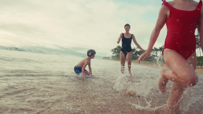 A mother and her two young children run and splash playfully in the shallow ocean waves on a tropical beach in slow motion