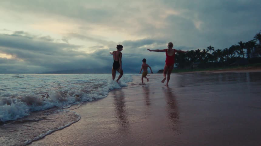 A mother, partner, and their young son run happily in slow motion along a tropical shoreline as waves wash over their feet at sunset