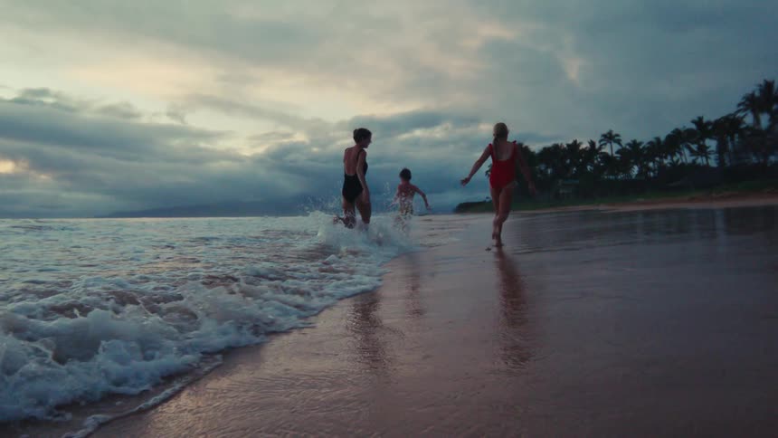 A mother, partner, and their young son run happily in slow motion along a tropical shoreline as waves wash over their feet at sunset