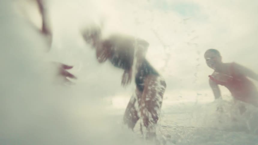 A joyful mother and her two children playfully splash each other in the ocean waves in an immersive, slow-motion shot