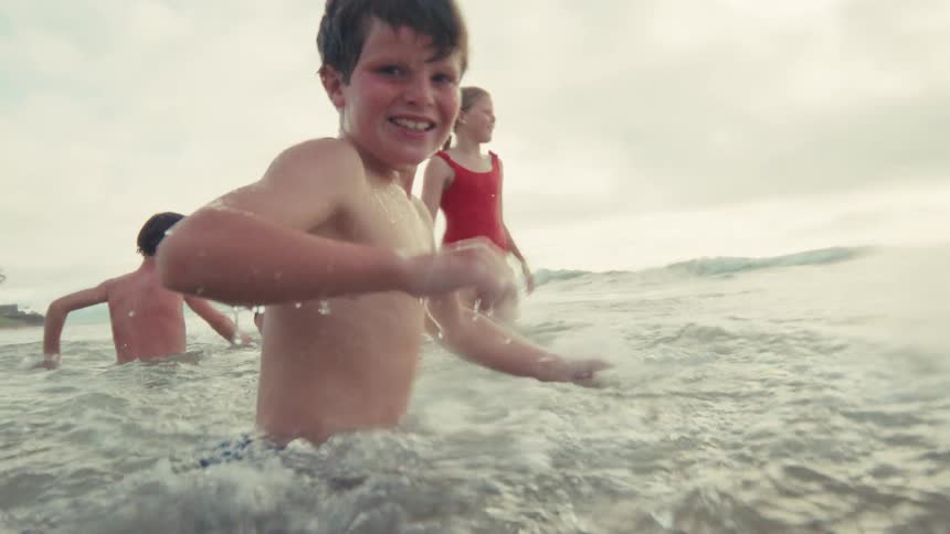 A happy young boy smiles and playfully splashes water at the camera while swimming in the ocean with his family in super slow motion