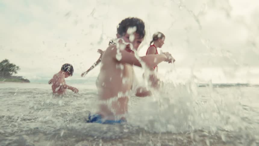 A happy young boy smiles and playfully splashes water at the camera while swimming in the ocean with his family in super slow motion