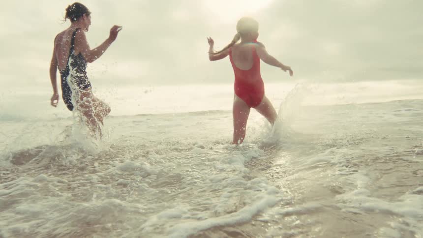 In a stunning slow-motion shot from behind, two women in swimsuits joyfully run into the ocean, splashing in the sunlit waves