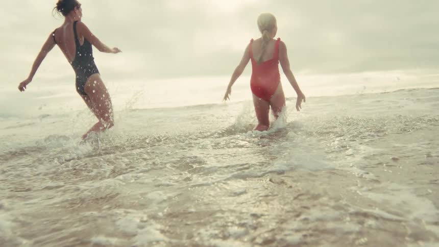 In a stunning slow-motion shot from behind, two women in swimsuits joyfully run into the ocean, splashing in the sunlit waves