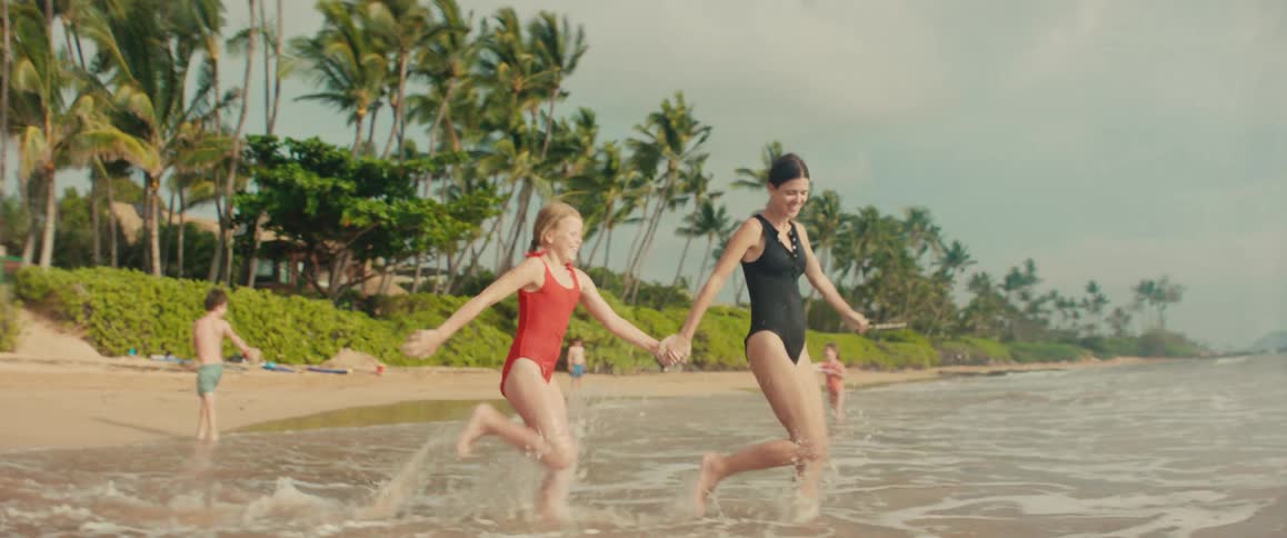 A smiling mother and her young daughter hold hands and run playfully through the shallow ocean waves in slow motion