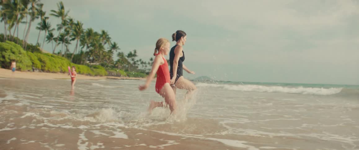 A smiling mother and her young daughter hold hands and run playfully through the shallow ocean waves in slow motion