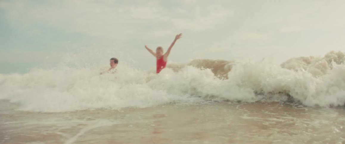 A large ocean wave crashes over a mother and her two children in slow motion as they play together at the beach