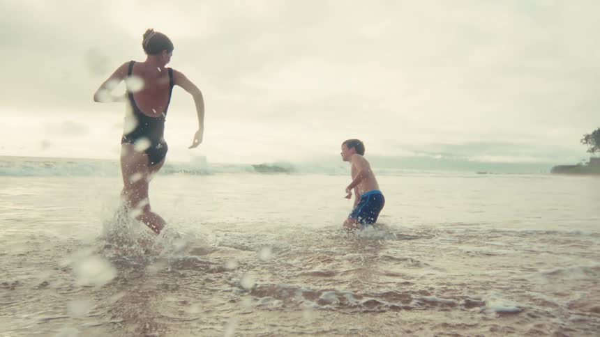 A mother and her young son joyfully run and splash in the shallow ocean waves in super slow motion
