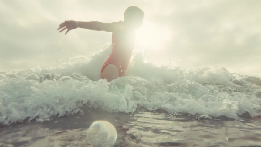 A young girl and her mother joyfully play in the ocean as a wave splashes over the camera in slow motion