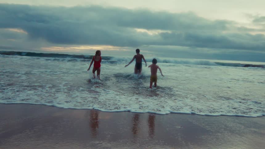 In slow motion, a woman and two children playfully run into the ocean waves on a beach during a cloudy evening