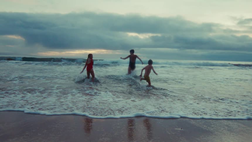 In slow motion, a woman and two children playfully run into the ocean waves on a beach during a cloudy evening