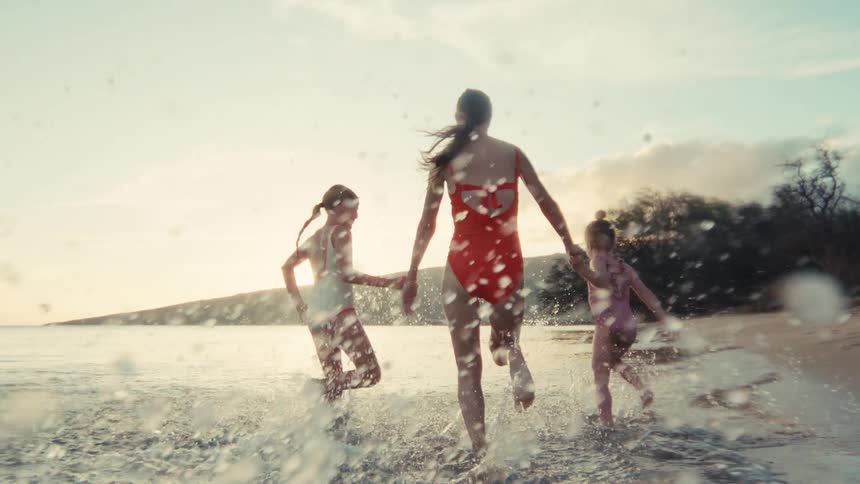 A mother and her two daughters joyfully run through the shallow ocean water at sunset, splashing in slow motion