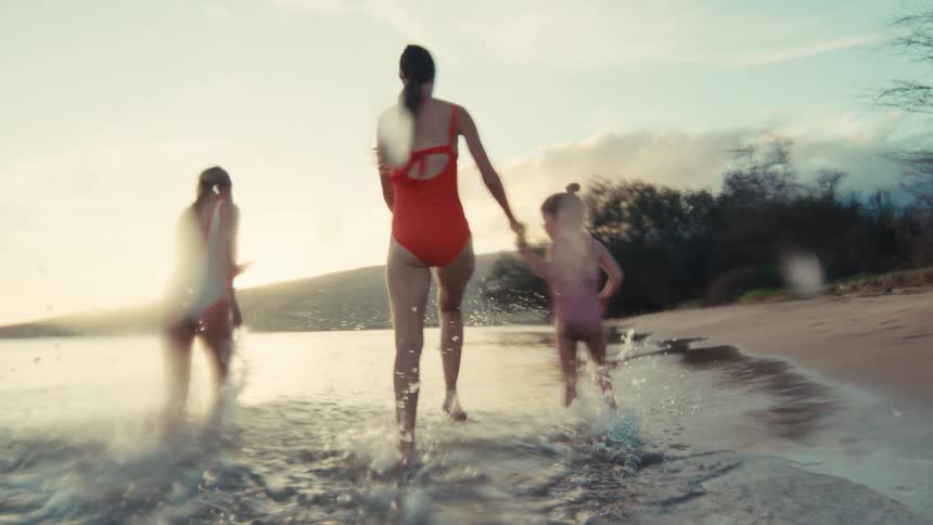 A mother and her two daughters joyfully run through the shallow ocean water at sunset, splashing in slow motion