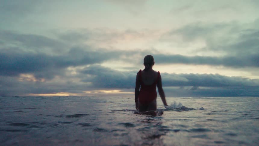 In slow motion, a woman in a vibrant red swimsuit walks into the serene ocean under a dramatic, cloudy sky at sunset