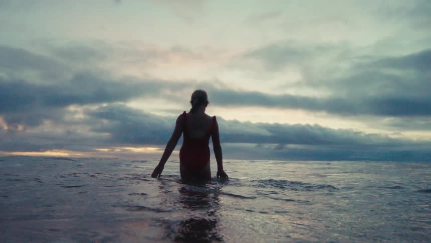 In slow motion, a woman in a vibrant red swimsuit walks into the serene ocean under a dramatic, cloudy sky at sunset