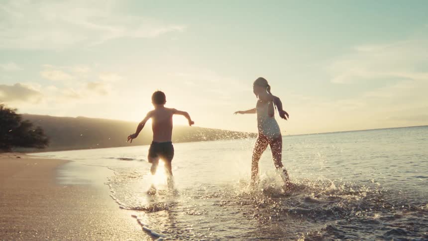 Two happy young kids run and splash in the shallow water along a sandy beach at sunset in slow motion