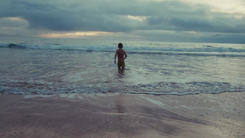 Slow motion shot of a young boy standing in the ocean waves as they wash ashore under a dramatic, cloudy sky
