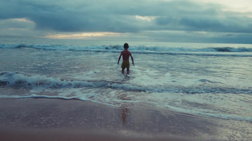 A lone young boy stands in the shallow waves of the ocean, peacefully contemplating the horizon under a moody, overcast sky in slow motion
