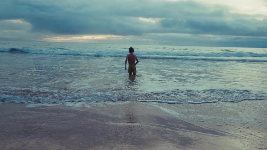 A lone young boy stands in the shallow waves of the ocean, peacefully contemplating the horizon under a moody, overcast sky in slow motion