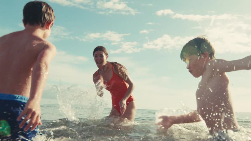 A mother and her two sons joyfully splash each other in the ocean during a sunny summer day in slow motion