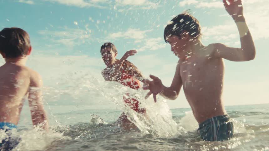 A mother and her two sons joyfully splash each other in the ocean during a sunny summer day in slow motion