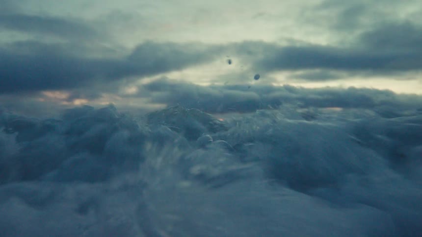 An immersive, low-angle slow-motion shot captures a powerful ocean wave crashing directly into the camera under a dramatic, cloudy sky at sunset